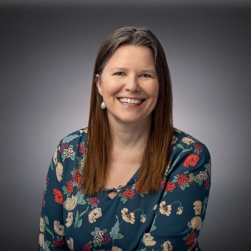 Smiling woman with long hair in a floral top, posed in front of a gray background for a professional headshot.