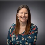 Smiling woman with long hair in a floral top, posed in front of a gray background for a professional headshot.