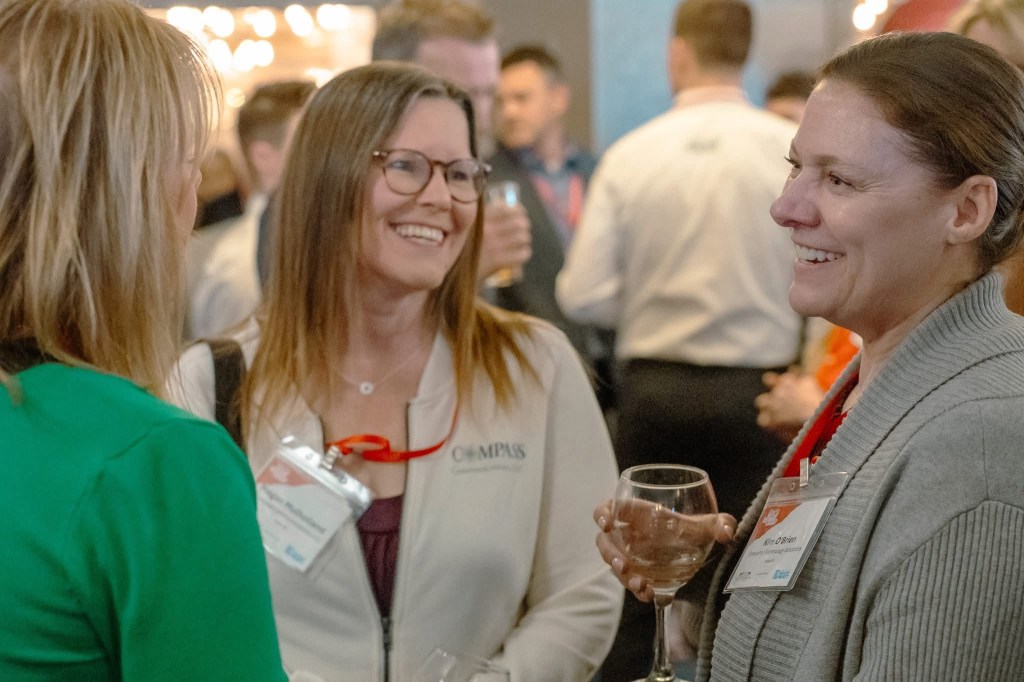 Three women smile and chat at a networking event; two wear name badges and hold drinks.
